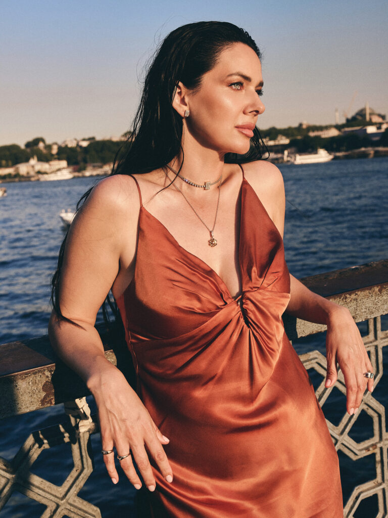 Jelena wearing layered Laleon necklaces and rings in a rust silk dress, leaning against a railing with the Istanbul skyline behind her