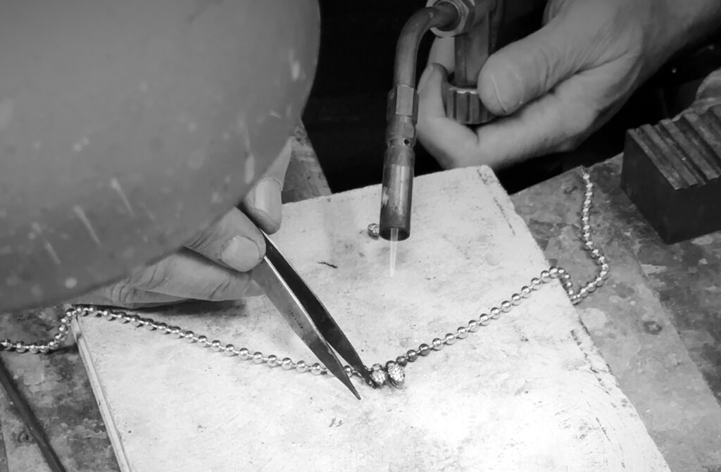 Artisan using a torch to solder a beaded gold chain by hand in the Laleon atelier, black and white detail shot